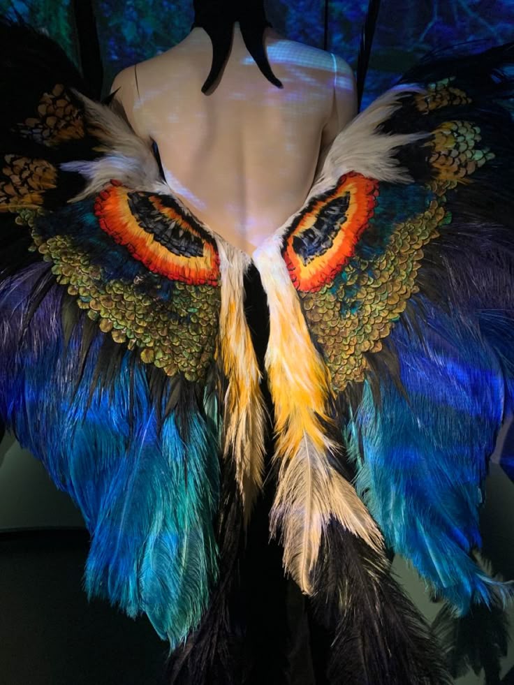 the image shows the back of a mannequin wearing a costume with large, colorful feathered wings. the wings are iridescent, with hues of blue, green, orange, and yellow. the costume also includes black hornlike adornments at the back of the neck. the feathers are meticulously arranged, creating a visually striking effect. the mannequin's bare skin contrasts with the texture and vibrancy of the feathers. the overall aesthetic is artistic and fantastical.