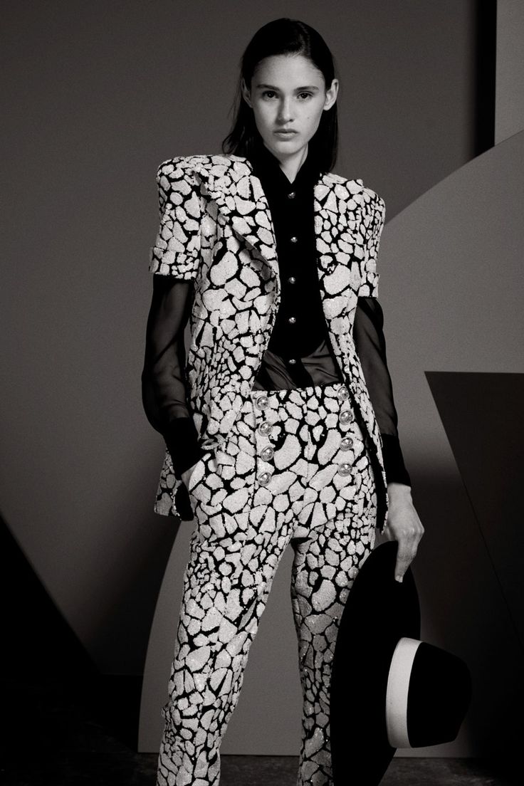 a black and white portrait of a fashion model wearing a stylish black and white outfit. she is wearing a blazer with a unique pattern, matching trousers, and a black hat. she is standing and posing in a studio setting.