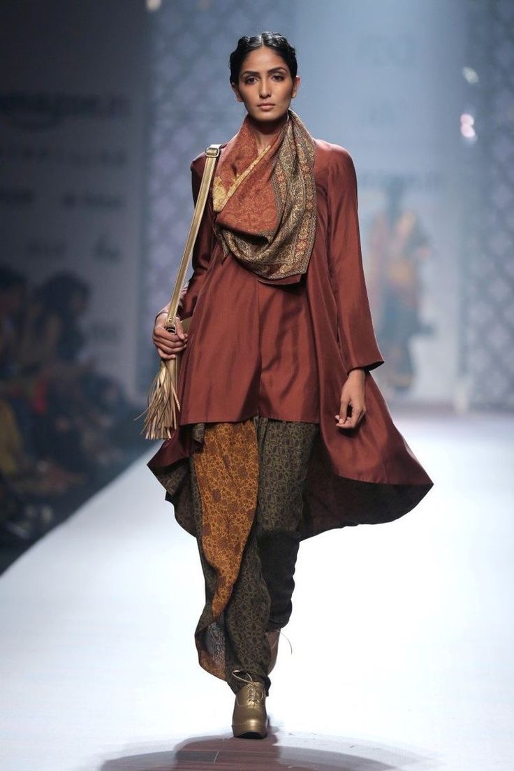 a model walks the runway wearing a stylish rustcolored asymmetrical tunic paired with printed dhoti pants.  she accessorizes with a patterned scarf, a gold handbag with tassels, and gold shoes. the outfit is a blend of traditional indian elements with a modern, western silhouette. the overall look is sophisticated and chic, suggesting an autumn/winter collection.  the background shows a blurredout audience and runway setting.