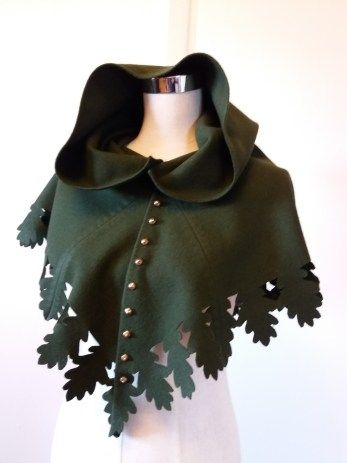 a green hooded cape with oak leaf cutouts along the bottom edge and fastened with buttons is displayed on a mannequin. the cape has a wide, draped hood and the leaf cutouts create a scalloped effect.