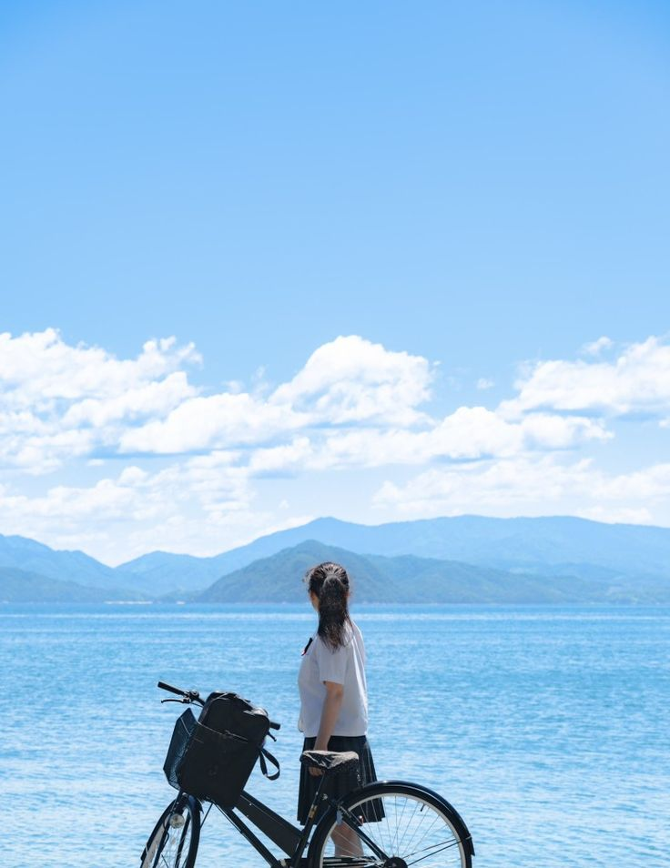 a young woman dressed in a school uniform stands with her bicycle on the shore, gazing out at the sea and distant mountains under a blue sky dotted with white clouds.