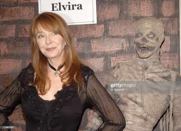 elvira, mistress of the dark, poses with a zombie beside a brick wall. she is wearing a black dress with a ruffled neckline and has a sweet smile on her face, while the zombie stares at the camera with a wide grin.  the name "elvira" is written on a sign above her head.  the image appears to have been taken at a studio.