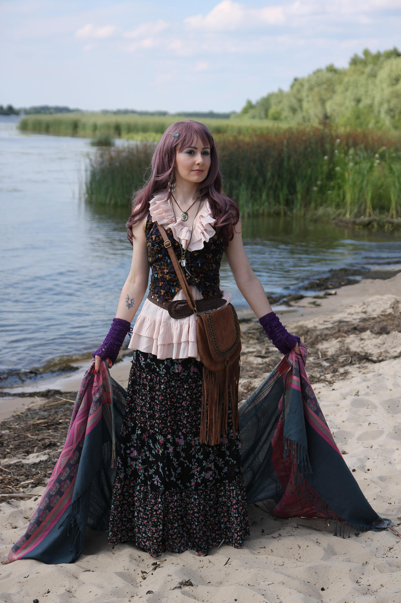 a young woman, possibly cosplaying or styled as a bohemian/gypsy, stands on a sandy beach by a river. she wears a long, floralprint maxi skirt, a ruffled pink and darkcolored top, and various accessories, including a fringed leather bag, bracelets, and a necklace. she is holding a large, darkcolored shawl or wrap. the setting is serene and natural, with a calm body of water and lush green vegetation in the background.  the overall aesthetic is vintageinspired and romantic.