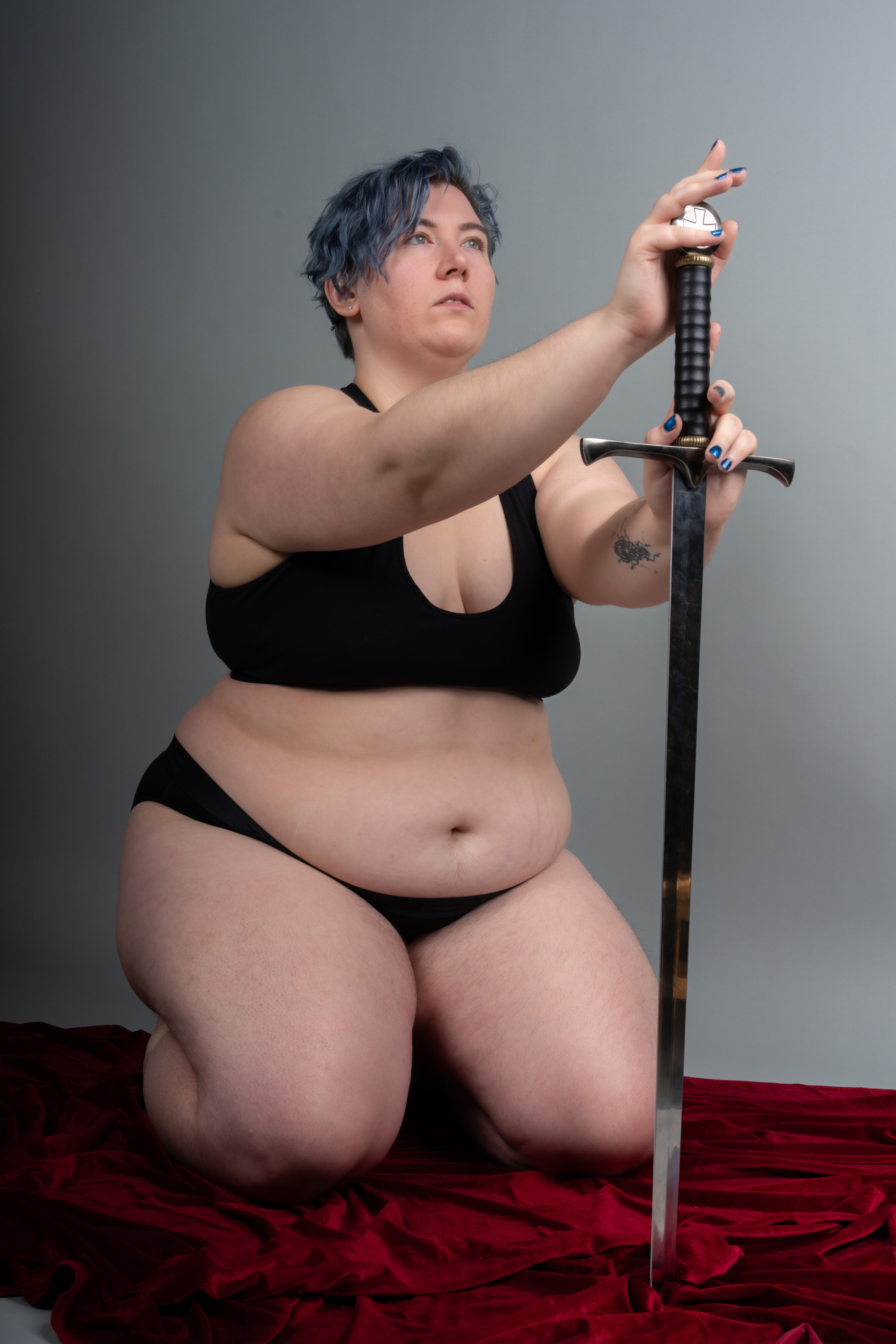 a woman with blue hair is kneeling on a red velvet cloth, wearing black lingerie and holding a sword. the background is a solid gray color.