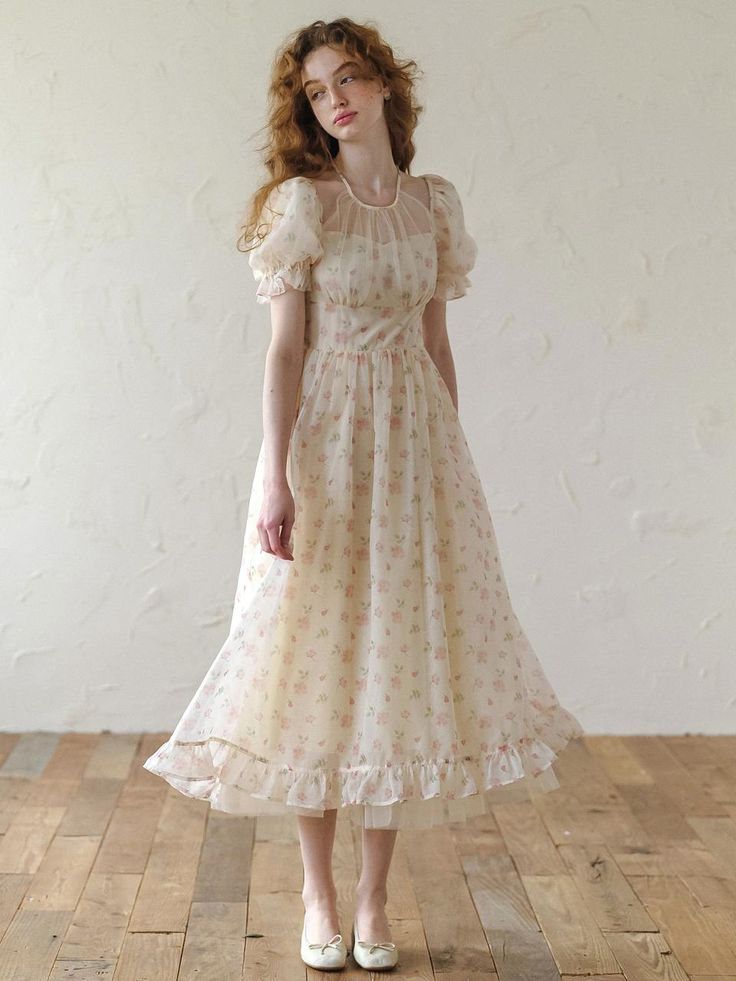 a young woman with long, flowing red hair models a vintageinspired, romantic dress. the dress is a light cream color with a pink floral print and features sheer puff sleeves, a ruffled hem, and a high neckline. she is posing in a room with a rustic wooden floor and white walls, illuminated by soft, natural light. the overall aesthetic is delicate, feminine, and dreamy.