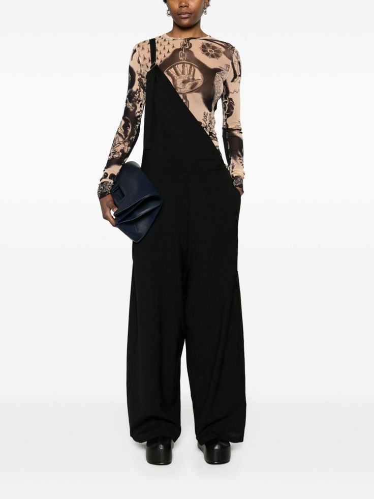 a young woman is standing in a studio, wearing a black overall jumpsuit with a seethrough longsleeved mesh top underneath. she is also wearing black shoes and carrying a small black bag.