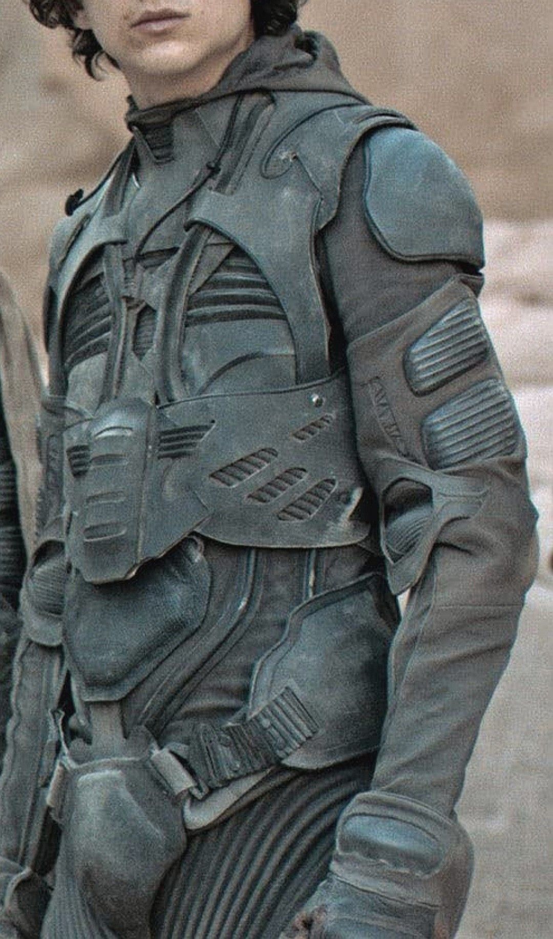 a closeup shot of a man wearing a futuristic, protective suit with a dark, textured fabric and armor plating. he is likely a character from a science fiction film or television show, possibly from the film dune.  the man appears to be in a desert environment. the suit's design is detailed and intricate, with a variety of textures and shapes.  the image evokes a sense of adventure, danger, and the unknown.