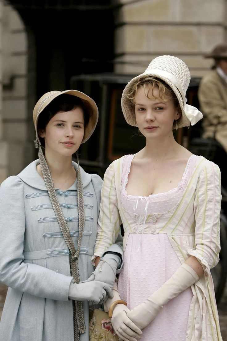 two young women, dressed in elegant 19thcentury style clothing, stand sidebyside.  the woman on the left wears a light blue coatdress and bonnet, while the woman on the right wears a light pink gown and a widebrimmed hat. both women are wearing long gloves and are holding hands. the setting appears to be an outdoor location, possibly a historical setting based on the architecture visible in the background.  the overall impression is one of friendship and historical context.