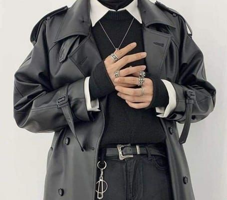 a person is wearing a stylish black outfit consisting of a black leather trench coat over a black sweater with a white collared shirt underneath. they are also wearing black jeans with a silverbuckled belt and a silver keychain hanging from the belt loop. the person is wearing a silver necklace and several silver rings on their fingers. the image focuses on the upper half of the body, highlighting the clothing and accessories.