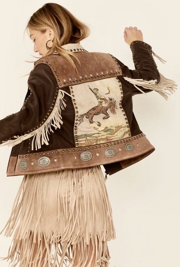 a back view of a woman's model wearing a brown and beige leather and suede jacket with fringe detailing and embroidery. the jacket features studs and metallic embellishments. the model is also wearing a matching beige fringe suede skirt. the style is bohemian, western, and country inspired. the image is taken on a white background.