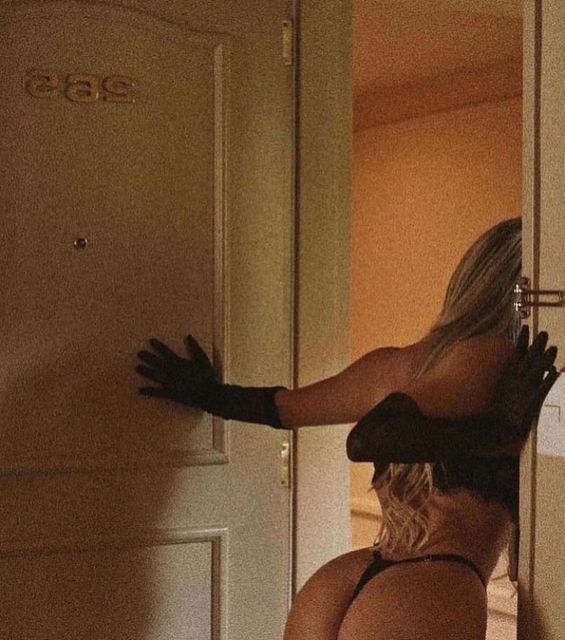 the image shows a blonde woman's back and backside, partially visible behind a hotel room door. she's wearing black lingerie and long black gloves.  she's leaning against and partially through the door, suggesting a sense of mystery and intrigue.  the room's lighting and color palette (muted oranges and browns) contributes to a retro or 70s aesthetic. the number "265" is visible on the door. the overall mood is sensual and suggestive.