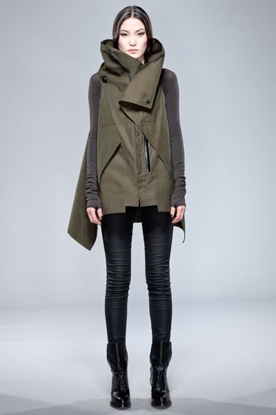 a young woman wearing a long dark green vest style coat with a high black collar and black pants, standing in a studio, looking toward the camera.