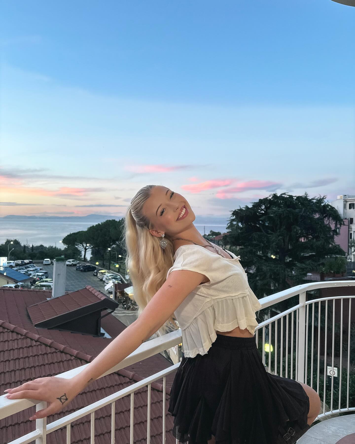 a blonde woman stands on a balcony wearing a white crop top and black skirt with her legs crossed. in the background, there is a sea and sunset view. the background also includes trees and buildings of a town.