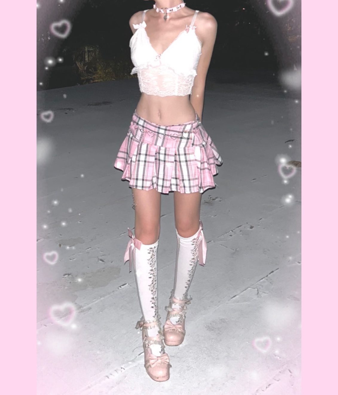 he image shows a young woman wearing a pink and white plaid pleated skirt, a white lace crop top, long white socks with pink bows, and pink platform shoes.  she is posing outdoors at night. the image is stylized with added hearts and a soft, pastel aesthetic.
