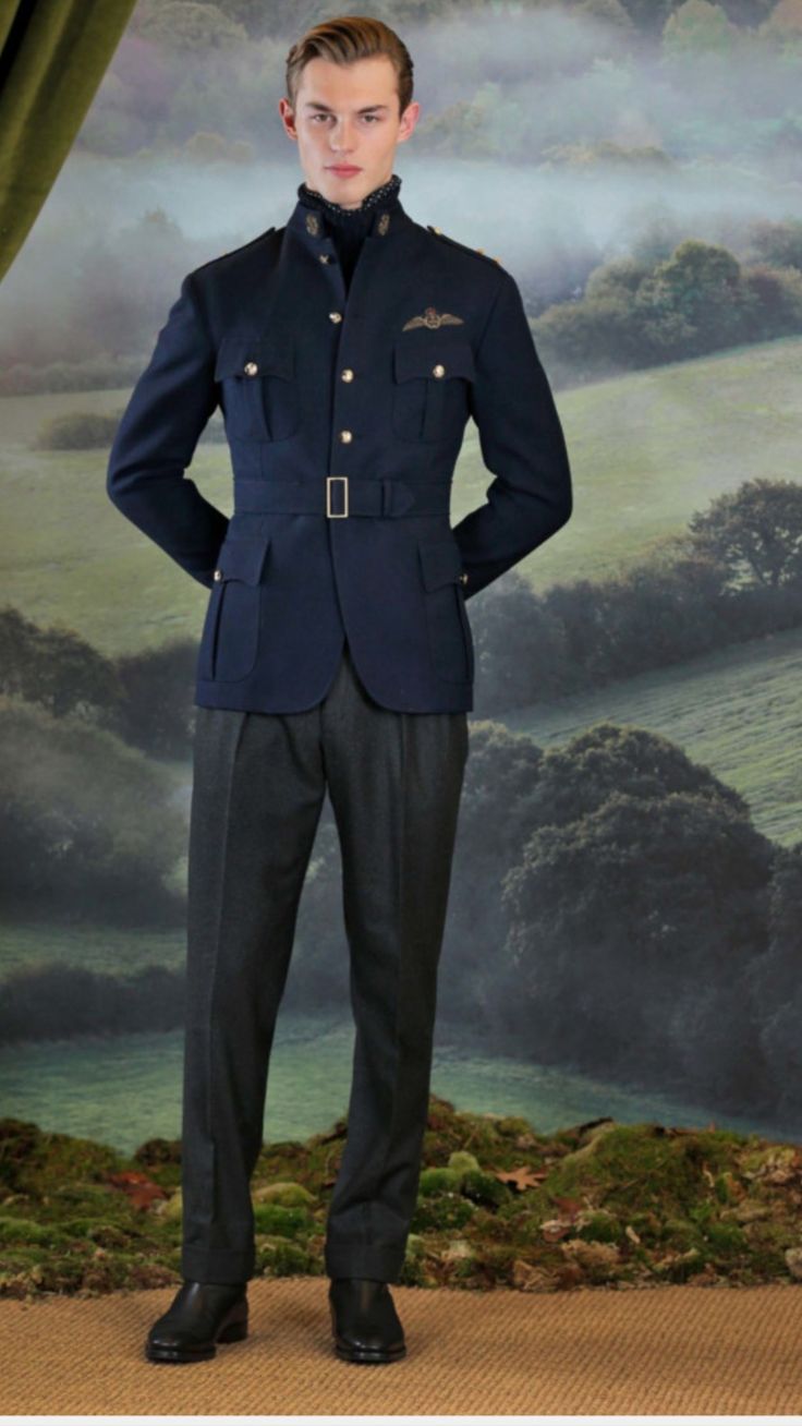 a male model is standing outdoors, wearing a stylish dark blue militarystyle jacket that resembles a royal air force pilot's uniform, complete with embroidered wings. the jacket has a belted waist and several buttons.  he is also wearing dark grey trousers and black shoes. the background is a picturesque landscape with rolling hills, greenery, and a slightly misty atmosphere.  the overall aesthetic is sophisticated and classic, suggesting a highfashion photoshoot.