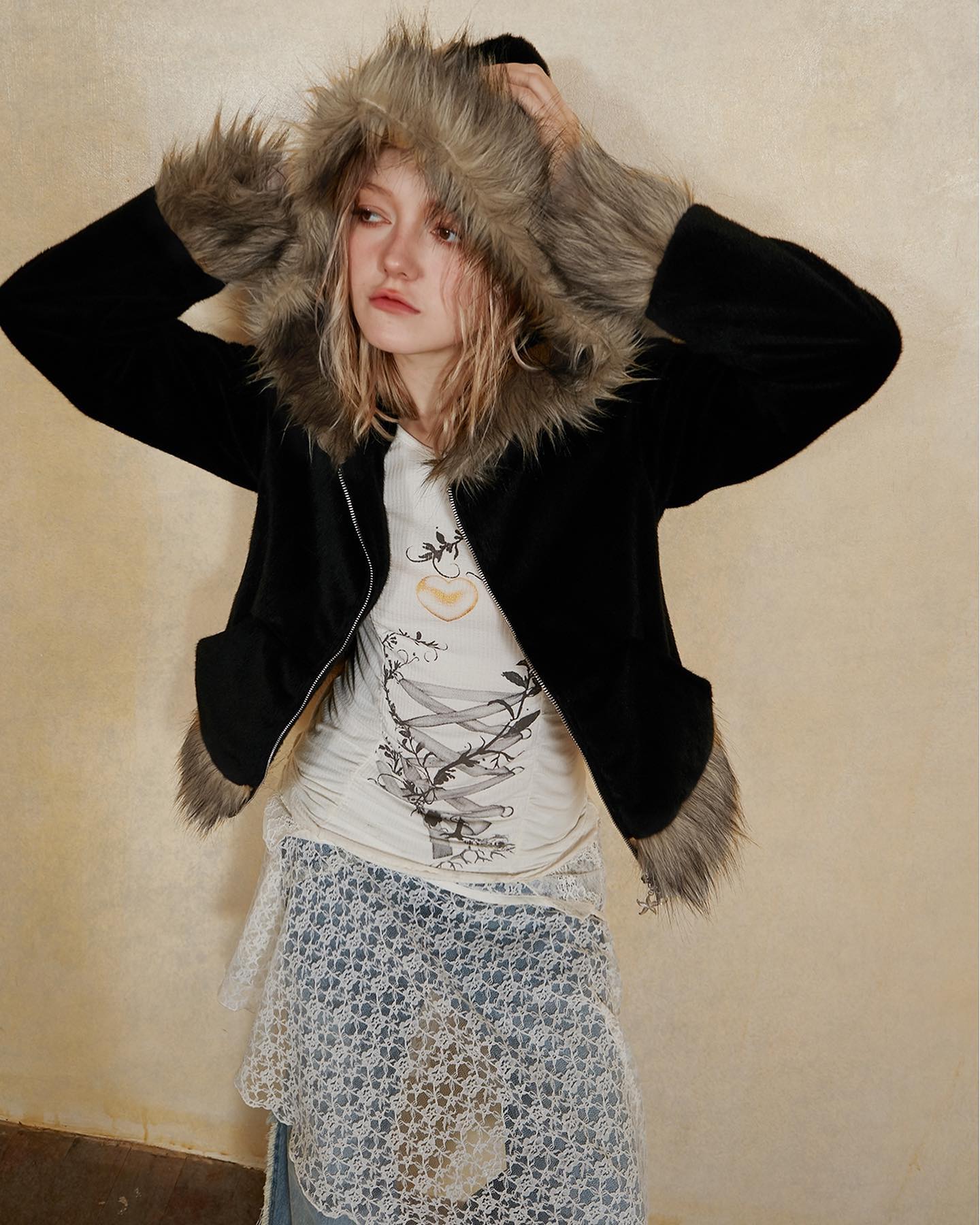 young woman with blonde hair is modeling a black hooded jacket with faux fur trim. the jacket has a zipper closure and is worn over an offwhite graphic tee and a lace skirt.  the model is posing against a beige background. the overall style is casual and slightly bohemian.