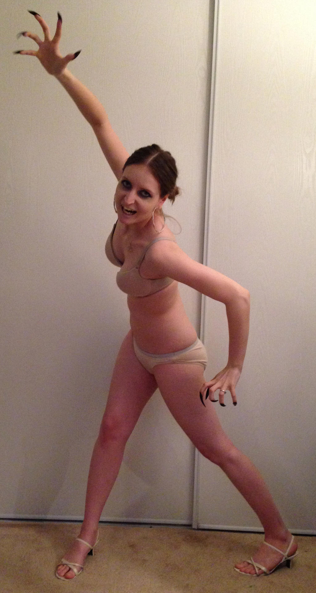 a young woman in her underwear is playfully posing in front of a white wall. she is wearing a beige bra and panties, and beige high heels. her nails are long and black. she has her arms up in the air and is smiling.