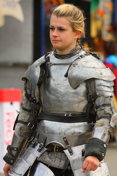 a woman is shown from the chest up wearing a full suit of plate armor. her blonde hair is pulled back in a loose style. the armor appears to be constructed from multiple pieces and includes shoulder plates, arm protection, a breastplate, and waist protection. she has a serious expression. in the background, other figures and objects can be seen, suggesting she is at a fair or festival. the armor looks worn and slightly tarnished.