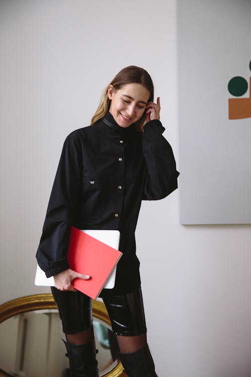 a young woman with long brown hair stands indoors, smiling, and holding a red and white laptop. she is wearing a black, buttondown shirt with a high collar, shiny black shorts, sheer black stockings, and tall black boots. a goldframed mirror is visible in the lower left corner, and a modern art painting hangs on the wall behind her. she has one hand raised to her hair.
