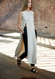 a woman is wearing a white dress with a slit up the front. the dress is paired with black pants and black sandals. she is walking on a wooden surface and looking straight ahead. the background is an outoffocus rustic wall. the woman's style is casual yet elegant.