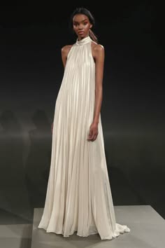 a woman is wearing a simple white halter neck gown. the gown is floorlength and pleated. the woman is standing on a black runway and is posing for a photograph. the background is dark and there is studio lighting. the model's hair is pulled back and she is wearing makeup.