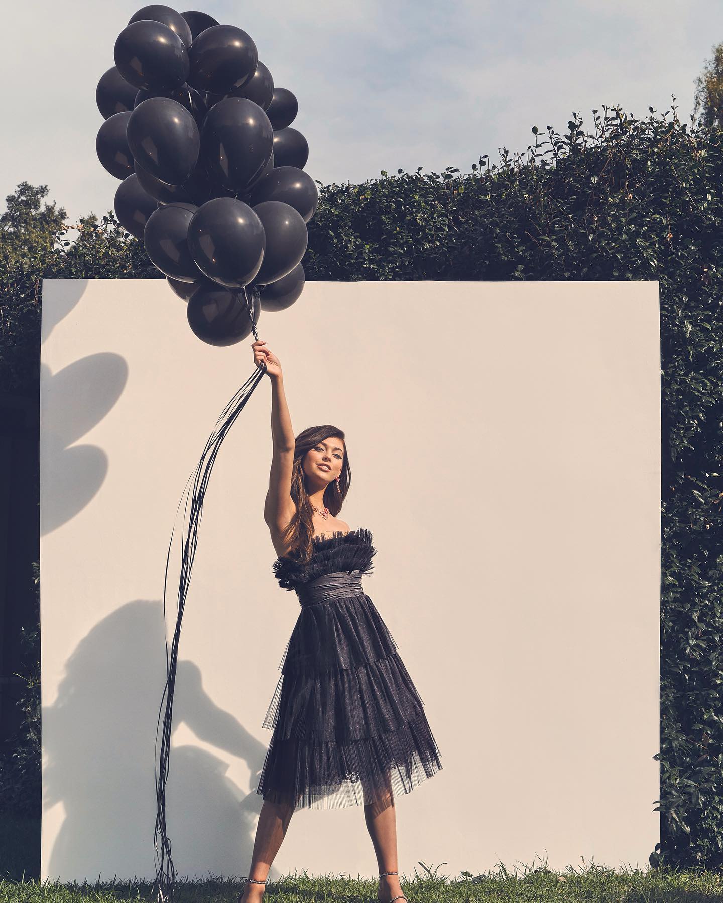 a woman in a black dress and heels stands outside holding a bunch of black balloons. she is in front of a white wall with a green hedge behind it. there is grass on the ground and trees in the background.