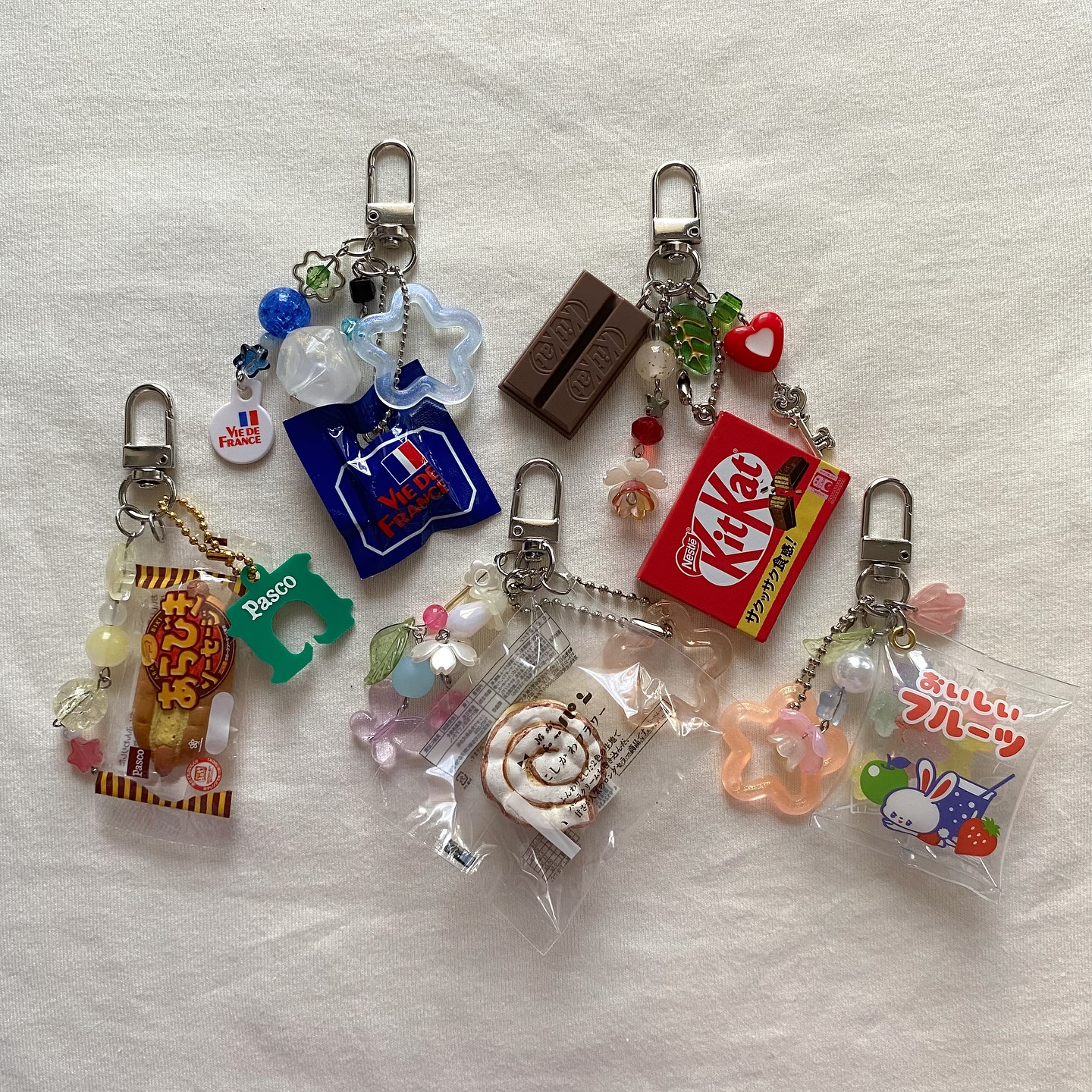 the image shows a collection of keychains, each featuring various charms related to food and sweets. there are charms depicting popular brands like pasco, vie de france, and kit kat, along with candy and chocolate representations. additionally, the keychains are adorned with beads and attached to clips.