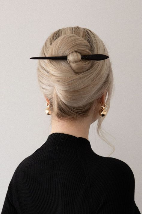 a woman with long blonde hair is wearing a black top. her hair is styled in a bun, with a black hair stick holding the bun in place. she is wearing a pair of gold earrings. the woman's back is facing the camera.