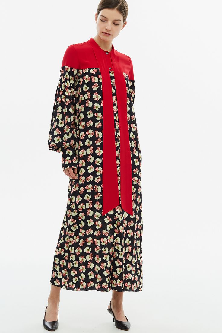 a woman is showcasing a long floral dress. the dress is predominantly black with a light beige and pink floral pattern. the top portion of the dress, including the collar and a center panel, is a bold red.  the dress has long sleeves and a relaxed fit. the woman is wearing black heels and has her hair pulled back. she appears to be posing for a fashion photograph against a simple white background.