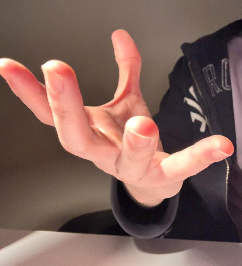 a closeup view of a right hand, reaching outward with fingers spread. the hand appears to be in a grasping or reaching motion. the person's sleeve is visible, suggesting they are wearing a darkcolored hoodie or jacket. the background is a neutral beige tone, typical of an indoor setting. the lighting suggests indoor lighting, possibly artificial light. the image is taken from an eyelevel perspective, capturing the hand in sharp focus.