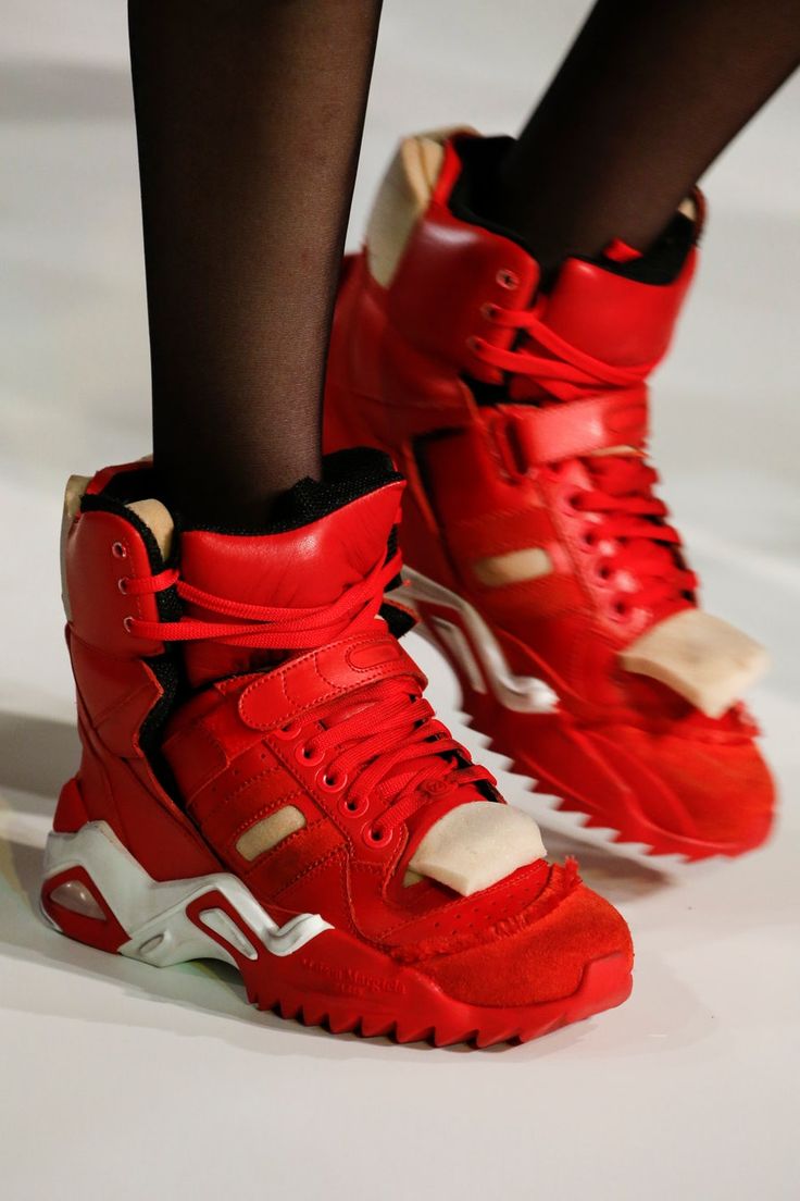 a model wears a pair of red high top sneakers.  the sneakers are a designer pair with white soles and laces. the model's other foot is wearing a similar pair of sneakers, but the laces are different. the sneakers are a statement piece of fashion and street wear.