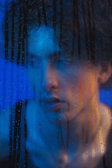 a closeup, blurry portrait of a young man's face seen through a raincovered window. the image is dark and moody, with the blue light illuminating the subject's face from behind the glass. the condensation on the window creates an abstract effect, obscuring details and adding a layer of mystery to the image. the subject's expression is pensive and contemplative.
