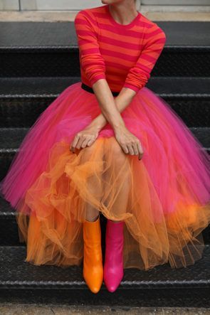 a woman wearing a bright red and orange striped sweater and a tulle skirt with a gradient of pink, orange, and yellow is sitting on stairs. she's posing with her legs crossed and her hands resting on her skirt. she's wearing pink and orange boots.  the skirt is very full and billowing.