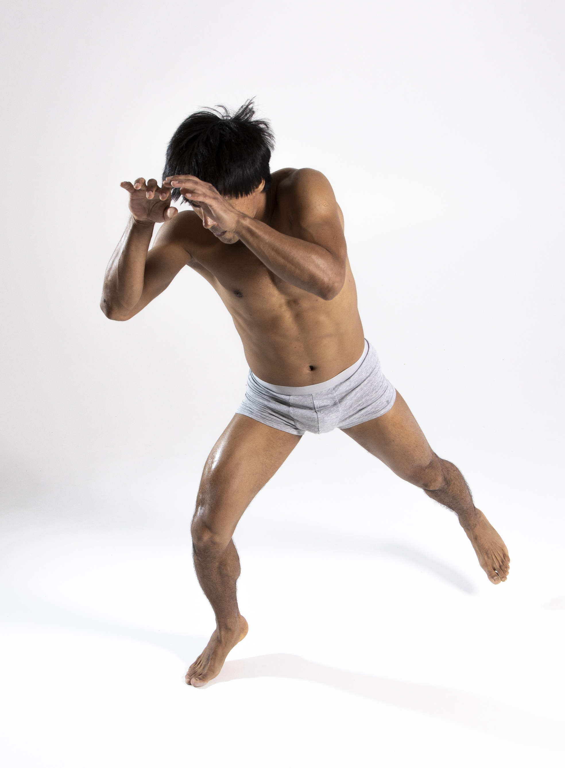 a shirtless, athletic man with dark hair is captured in a full body studio shot. he is wearing boxer briefs and is bare feet. he is in a defensive posture, covering his face with his hands and looking down, suggesting fear or anxiety. his body is toned, and he seems to be dodging or evading something. the background is plain and white.