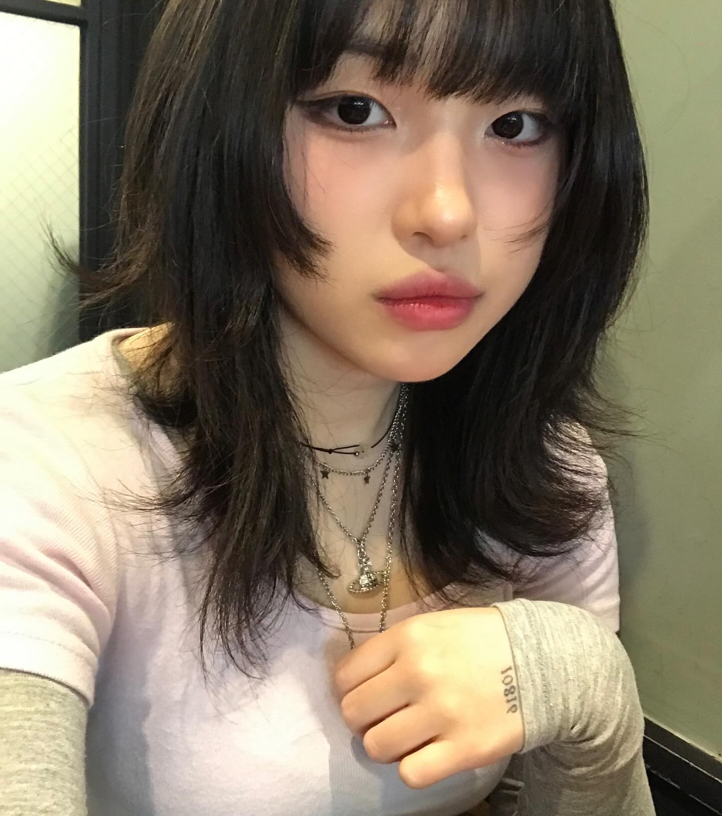 a  woman with a stylish wolf cut hairstyle takes a selfie. she has layered necklaces, rings on her fingers, and a small tattoo on her hand.  she is indoors and the image has soft lighting.