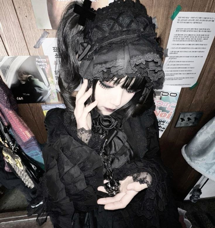 a closeup, slightly lowangle shot of a young woman dressed in elaborate gothic lolita fashion. she wears a black bonnet with lace detailing, a black dress with ruffles and lace accents, and a long black necklace with a large pendant. her dark hair is styled in pigtails, and she has bangs framing her face. she's holding a small object in her hands. the background is a cluttered wall with various posters and items, creating a grunge aesthetic. the overall mood is dark, mysterious, and stylish, typical of alternative fashion subcultures. the focus is on the woman and her clothing, showcasing the intricate details of the gothic lolita style.