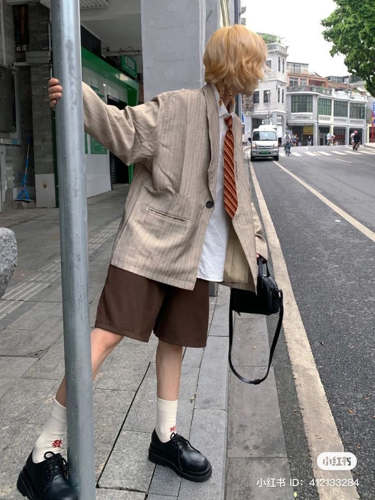 a young blonde woman wearing a striped blazer, brown shorts and black shoes poses on the sidewalk in a city setting. she is holding a black purse and is looking away from the camera.