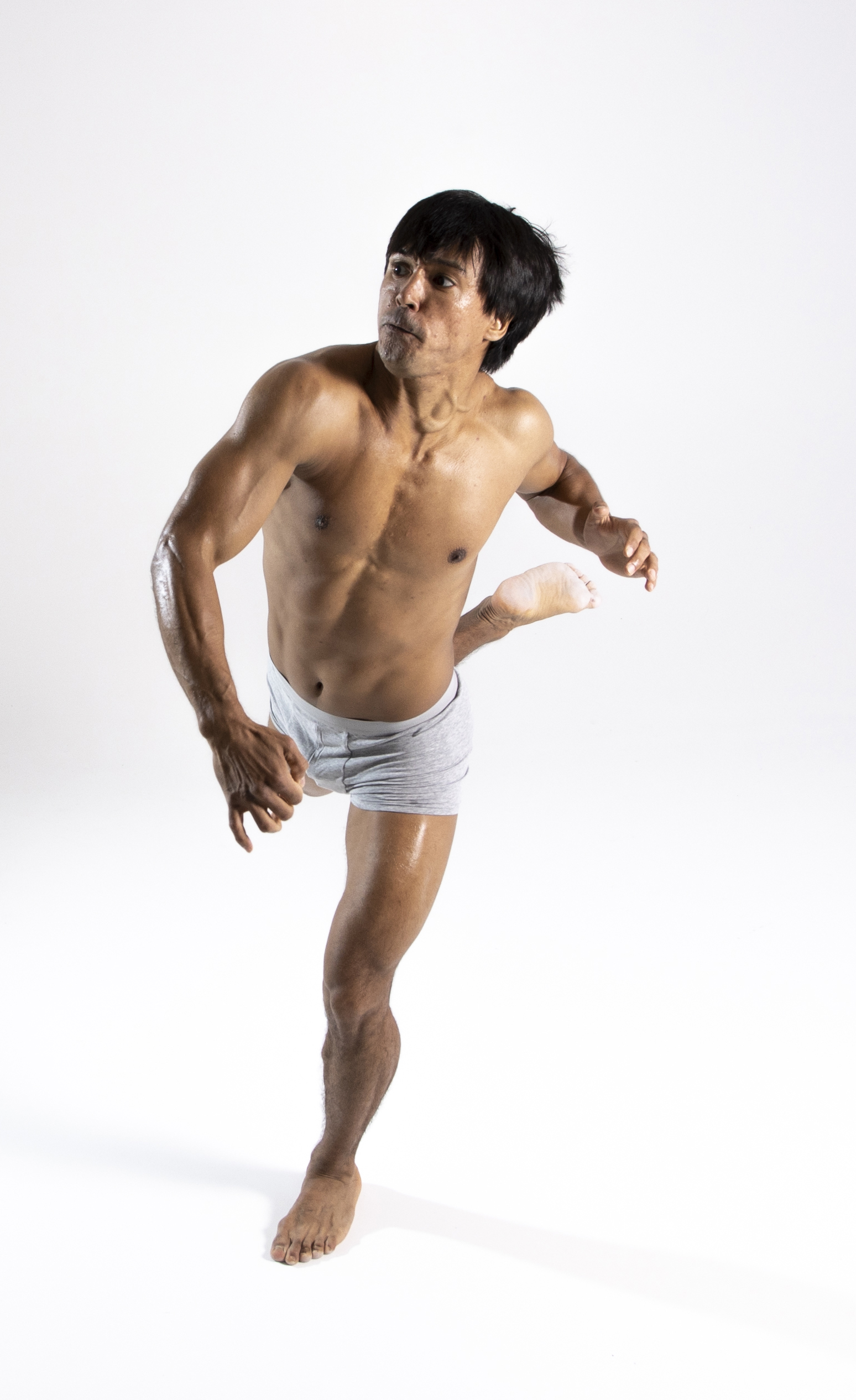 a shirtless, muscular asian man is posing against a white background in a dancelike position. he is wearing gray shorts and is barefoot. one leg is raised behind him. he is looking away from the camera.