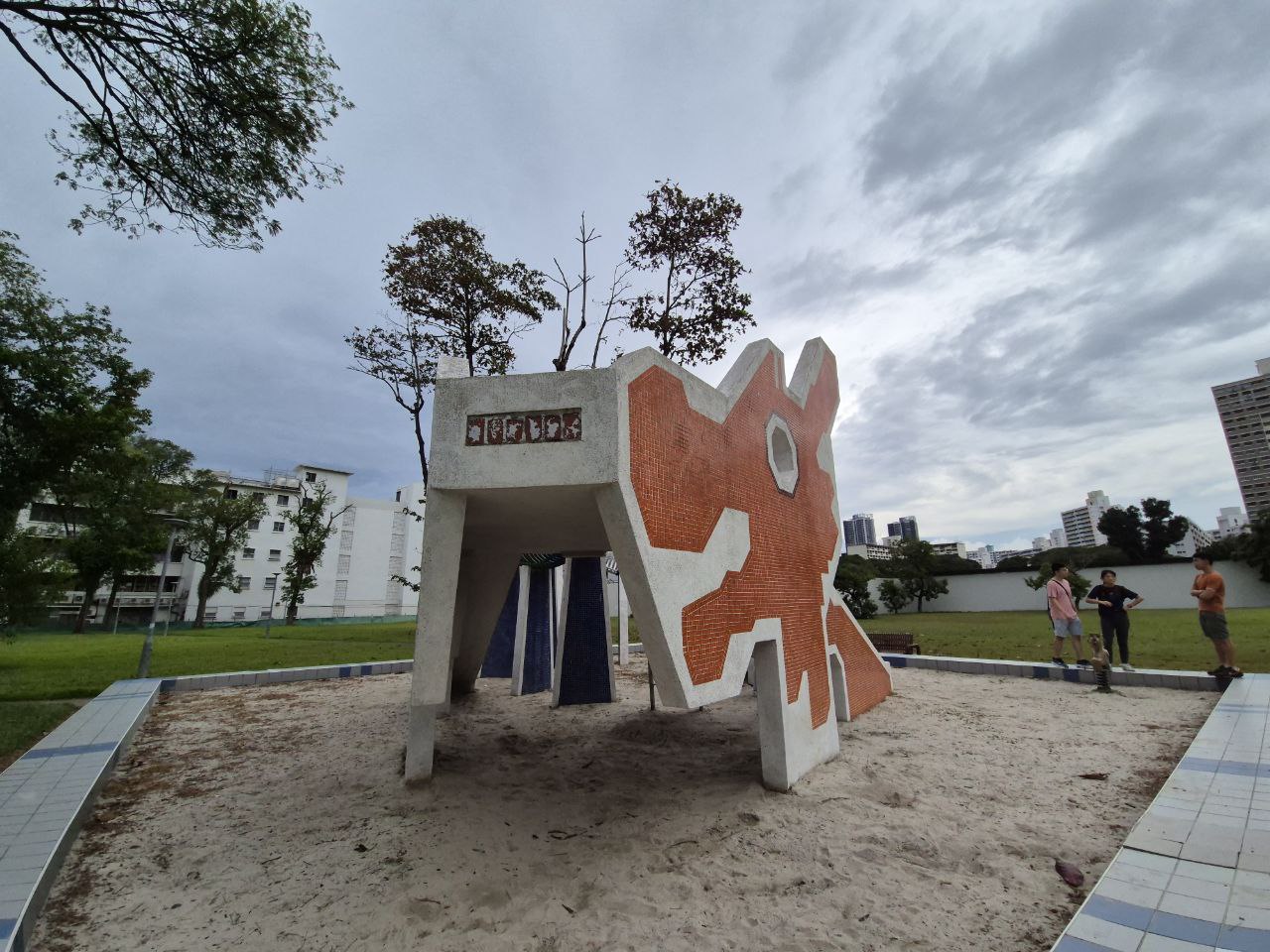 a concrete playground structure shaped like a large abstract sculpture with orange and white geometric designs is featured in this photo. the sculpture has multiple platforms, a climbing wall, and a slide.  the structure is situated in a grassy park with a small sandbox area in front of it. there are trees and buildings in the background and a few people are visible playing in the distance.  the sky is partly cloudy.