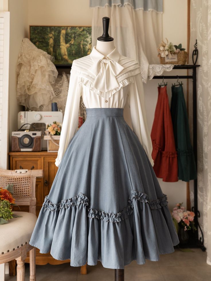 a fulllength view of a mannequin dressed in a classic lolita fashion outfit. the outfit features a flowing, slate blue skirt with layers of ruffles and a crisp white blouse with a ruffled collar and a bow. the mannequin is positioned slightly to the left of the frame, in a room with white walls and a wooden floor. the background is decorated with a white curtain and a wooden rack holding clothing items in warm colors. the lighting is bright and artificial, creating a clean and elegant atmosphere.