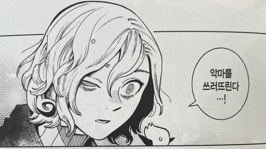 the image is a closeup panel from a manga or anime comic.  it features a young character with wavy, shoulderlength hair. the character's face is partially visible, showing a smirk or slight sneer with one eye partially closed. their expression is slightly mischievous. a speech bubble above them contains korean text. the art style is typical of manga/anime, with clear linework and shading. the overall tone is somewhat playful and slightly sinister.