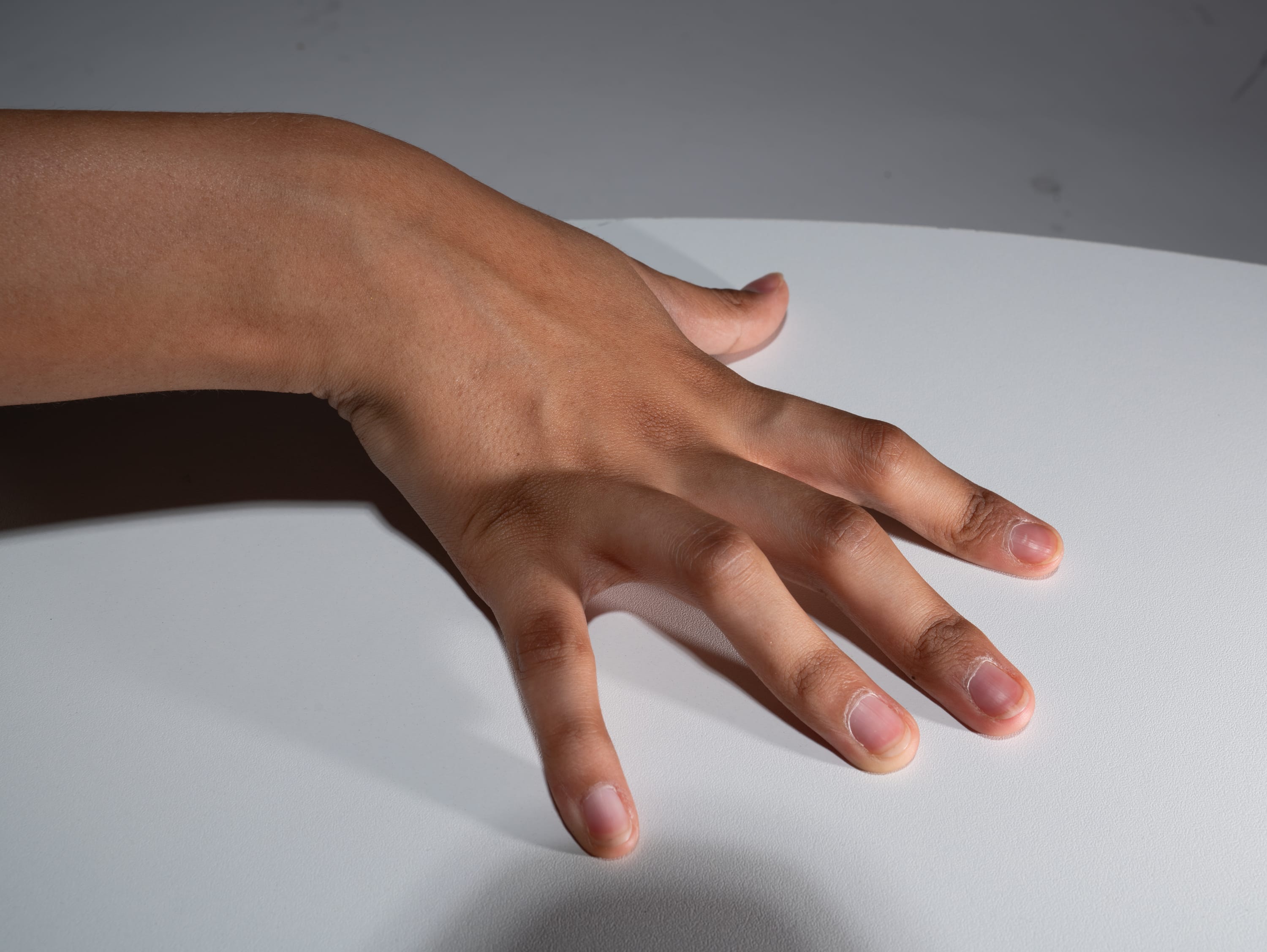 a closeup image of a hand resting on a white table. the hand is slightly spread, with the fingers extended. the lighting is soft and diffused, highlighting the details of the skin and nails. the image is taken from a closeup perspective, emphasizing the texture and form of the hand.