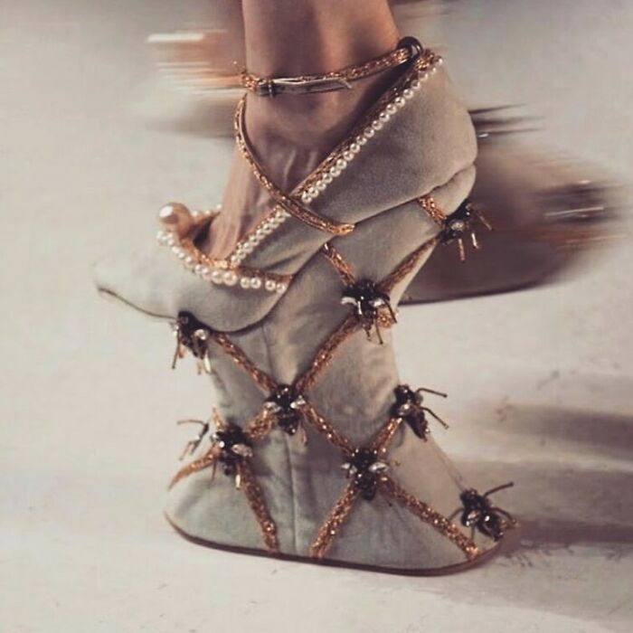 a closeup shot showcases a unique highheeled shoe adorned with intricate detailing. the shoe is primarily a muted color, embellished with gold trim forming a crisscross pattern. decorative bug accents are placed at the intersections of the gold trim. a string of pearls lines the top edge of the shoe. an ankle strap completes the design. the foot is seen partially inside the shoe.