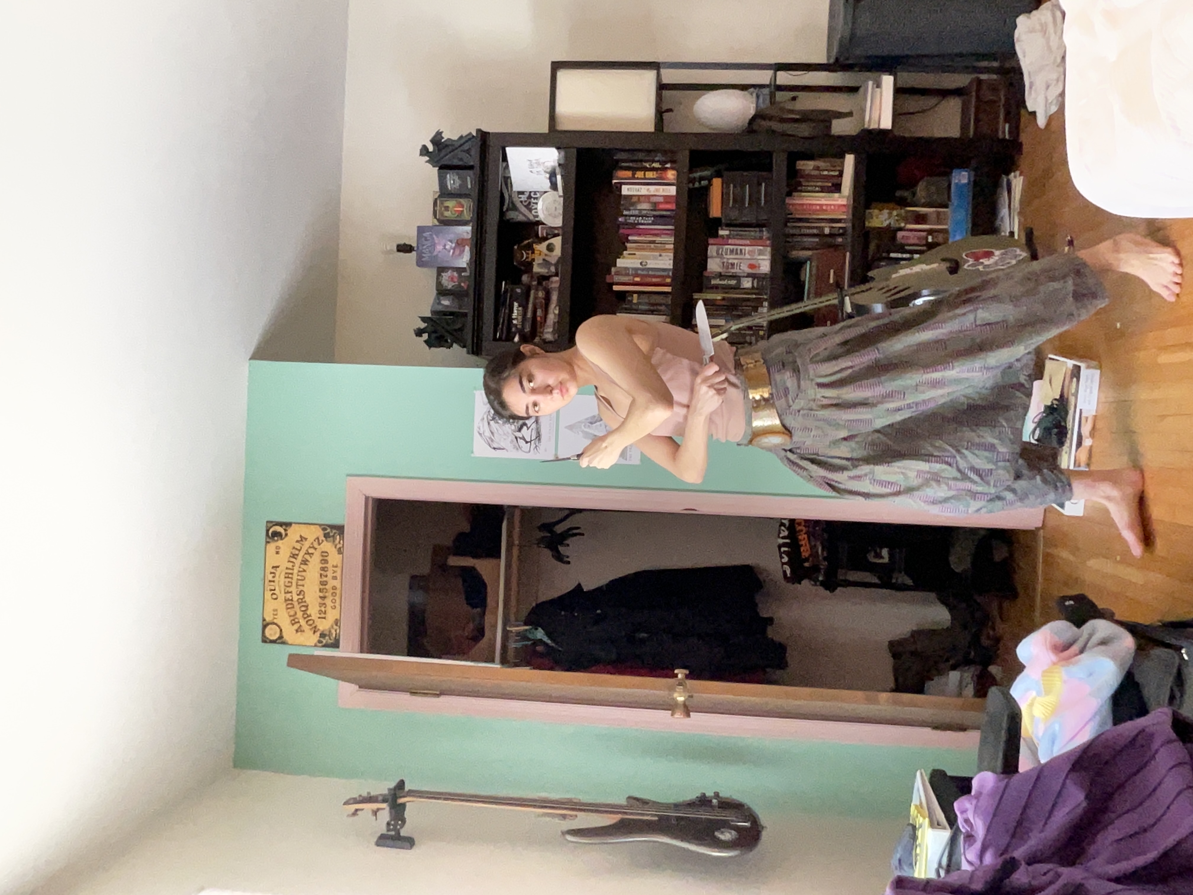 a young woman is standing in her bedroom, holding a knife in a selfdefense pose. she is wearing loosefitting, patterned harem pants and a gold belt.  the room is eclectically decorated, with a bookshelf full of books, a guitar leaning against the wall, and an ouija board hanging on the door.  she is in a martial arts or selfdefense stance.