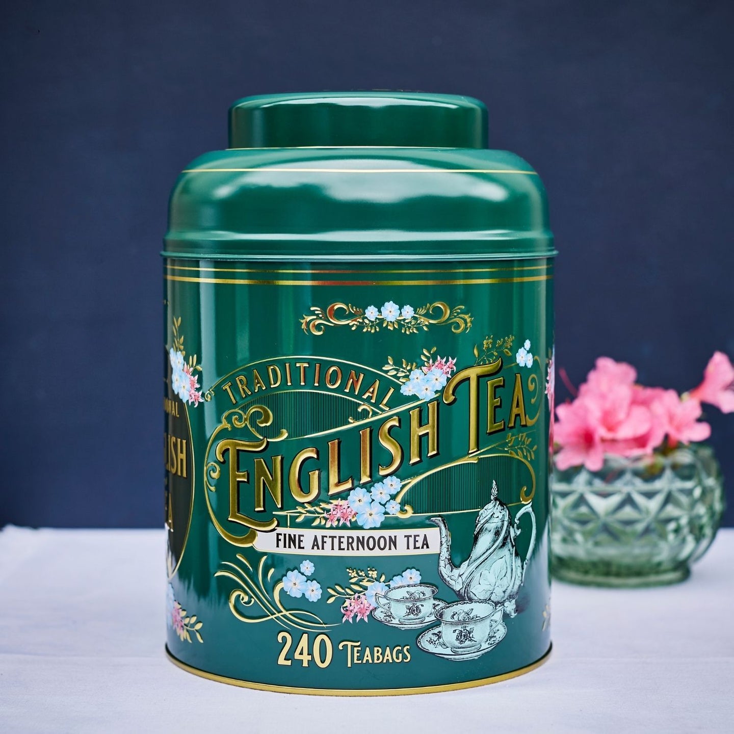 the image shows a cylindrical green tin canister of traditional english tea.  the canister is prominently labeled with gold lettering proclaiming "traditional english tea" and "fine afternoon tea," along with an illustration of a teapot, teacups, and saucers. the design incorporates a vintagestyle floral pattern. the tin sits on a white surface, and a vase of pink flowers is visible in the background. the overall impression is one of elegance and classic british teadrinking culture.  the label indicates it contains 240 teabags.