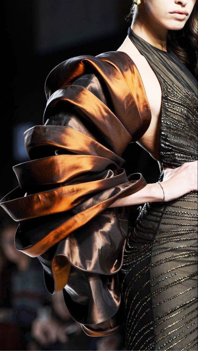 a woman on a runway is wearing an evening gown that has a dramatic sleeve. the sleeve is constructed out of a bronze colored, shiny fabric and resembles rounded, tiered bands. the dress has a deep halter cut and black embellishments on the front.