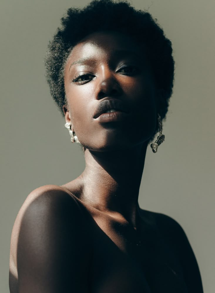 a portrait of a darkskinned woman with short, dark hair. she is wearing earrings and her shoulders are bare. the lighting is dramatic, with strong shadows and highlights. she is looking directly at the camera with a serious expression. the photo is likely taken indoors.