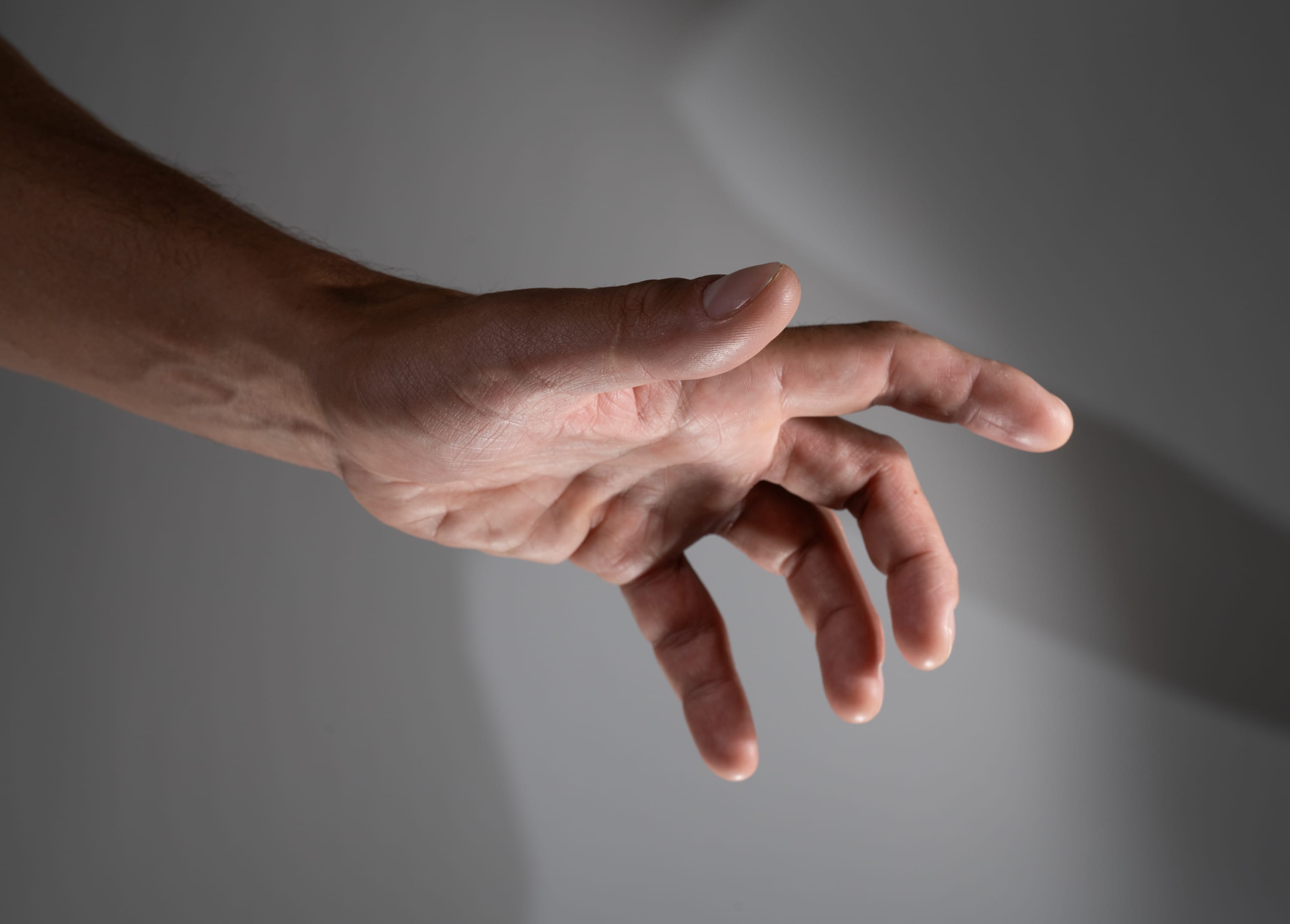 a male's hand is extended towards the viewer, fingers spread slightly. the hand is lit with soft, natural light that creates subtle shadows on the fingers and palm. the background is a neutral gray, drawing attention to the hand's gesture.