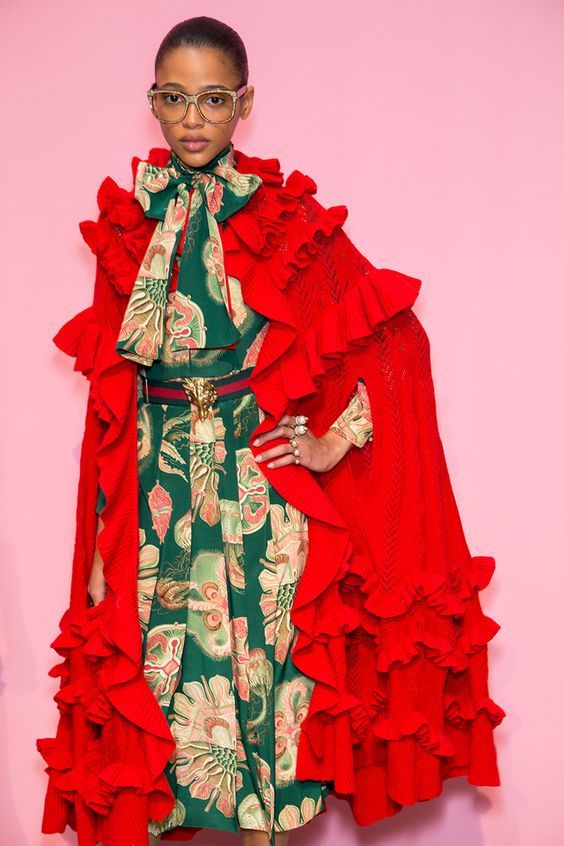 a fashion model is showcasing a striking outfit against a pink backdrop. the ensemble features a vibrant green floral dress adorned with intricate detailing. over this, a dramatic red cape with layered ruffles adds a bold contrast and texture. the model's accessories include stylish glasses, a gucci belt with a lion head buckle, and delicate rings. the overall presentation exudes elegance and sophistication, typical of a highfashion runway setting. the image effectively captures the details and impact of the clothing and accessories.