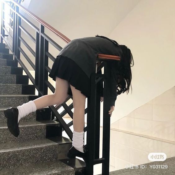 a person in a school uniform is leaning over a railing on a set of stairs. the person is wearing a dark skirt, black shoes, and white socks. the railing is black and the stairs are grey. the person's hair is dark and covers their face. the background is a white wall.